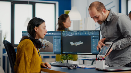 Industrial designer holding tablet discussing with woman engineer while working in CAD program, designing new components, comparing 3D prototype with project from notepad, using pc with two monitors