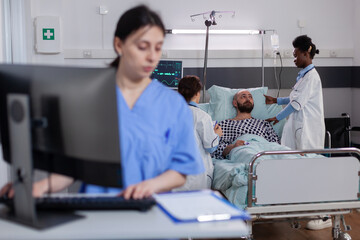 Front view of asisstant typing medical expertise on computer while in background black practitioner doctor discussing with sick man healthcare treatment. Hospitalized patient having respiratory