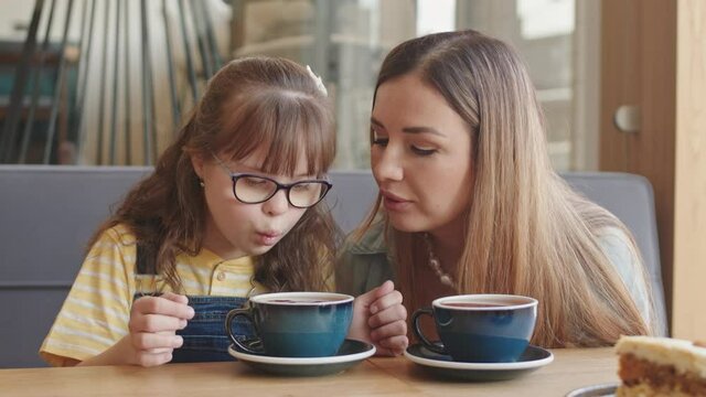 Medium slowmo of young Caucasian woman and her 11-year-old daughter with down syndrome blowing on cup of hot tea sitting at table in cozy cafe