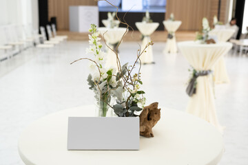 Tent cards placed on a tall table covered with white cloth decorated with twigs and white flowers at a wedding banquet in a hotel.
