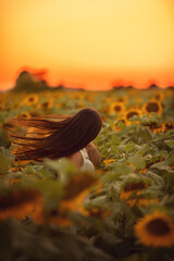 A happy, beautiful young girl with long flying hair in a straw hat is standing in a large field of sunflowers in the rays of the setting sun. Summer time. Back view. Selective focus