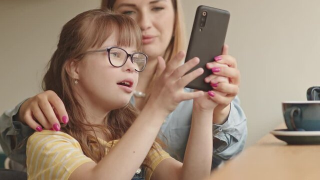 Waist-up shot of pretty elementary age girl with down syndrome holding smartphone and learning to take pictures while her affectionate mother helping her spending time together in cozy cafe