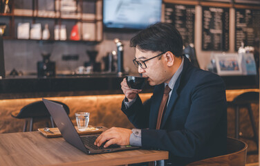 Asian businessman wearing suite and eyeglasses drink coffee , typing notebook for work at cafe restaurant. 