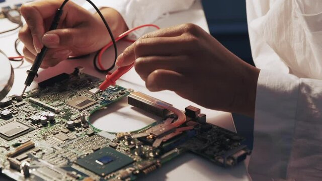 Close up of engineer's hands using multimeter tester to check motherboard in laboratory. The specialist is in white robe