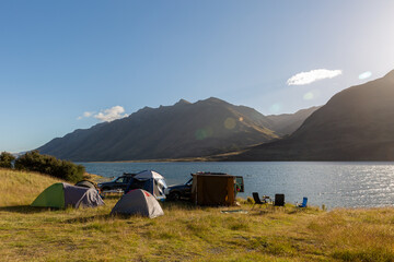 A photo of a group of tents put up by the lake. Three tourist chairs and two unrecognizable cars standing sideways behind the tents. Mavora Lake and Livingstone Mountains in the background. © Anna J. Photography