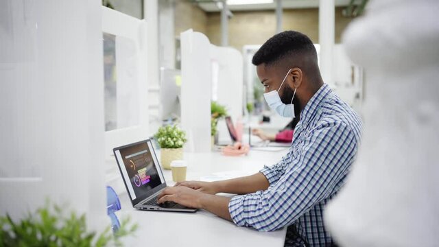 Focused black man with face mask sitting in his desk and using his laptop. Office concept.