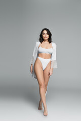 Fototapeta premium Sexy barefoot woman in underwear and silk robe walking on grey background