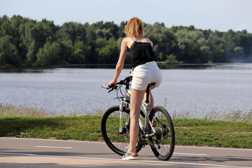 Fototapeta premium Slim redhead girl in shorts stopped with bicycle on river and forest background. Cycling and summer leisure