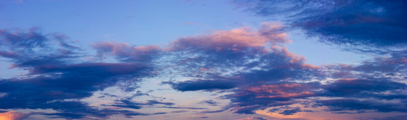 Panorama of blue sky, sunset and dark dramatic clouds illuminated by the orange sun.