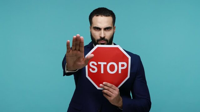 Serious bossy bearded male holding and showing red stop traffic sign, warning and asking to be attentive, speed limit, wearing black official style suit. Indoor studio shot isolated on blue background