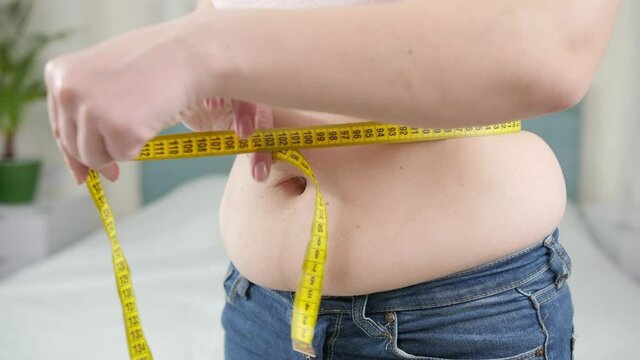 Closeup of obese woman measuring her big belly with yellow measuring tape. Concept of dieting, unhealthy lifestyle, overweight and obesity