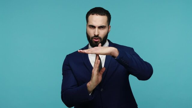 Exhausted serious young brunette attractive man in formal suit showing time out hand gesture and begging to remove deadline, needs break. Indoor studio shot isolated on blue background.