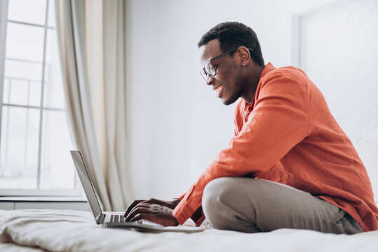 Smiling African-American man freelancer in bright orange shirt works on modern laptop on comfortable bed at home side view