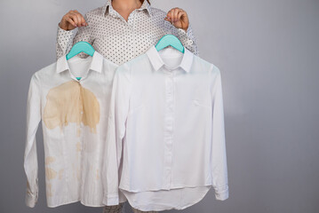 A woman compares two white shirts before and after washing. The girl is holding one blouse, clean and ironed, and the other, dirty with coffee stains