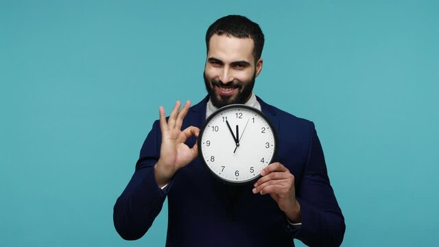 Brunette young bearded man in official style suit holding big wall clock and looking at camera with optimism, being happy finishing work in time. Indoor studio shot isolated over blue background.