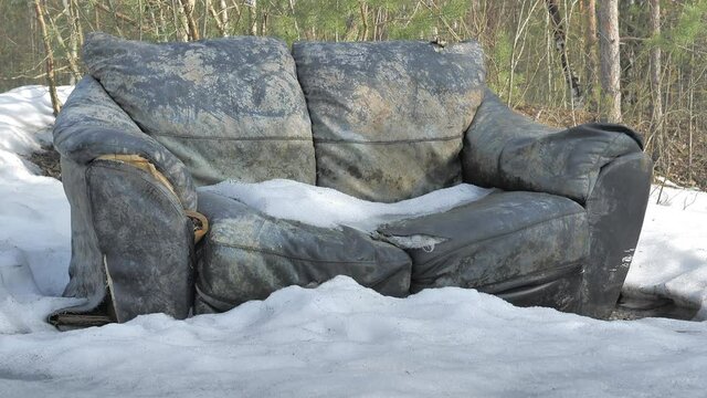 Closeup shot of a couch in a forest during winter. Polluted environment. Trash left in the beautiful nature by people. Camera slowly tilting upwards.