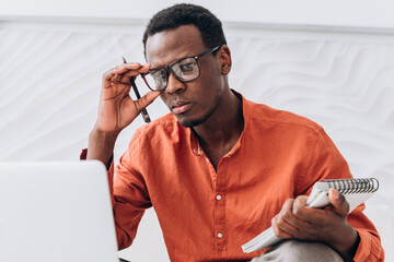 Serious African-American businessman with pen and paper notebook adjusts glasses looking at modern...