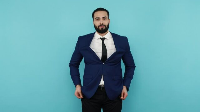 No cash, need financial support. Poor bearded businessman showing empty pockets inside out, having no money, debt problems and bankruptcy. Indoor studio shot isolated on blue background.
