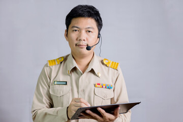 A male Thai government teacher in a headset with headphones standing holding a tablet and looking at the camera on gray background, online teaching concept