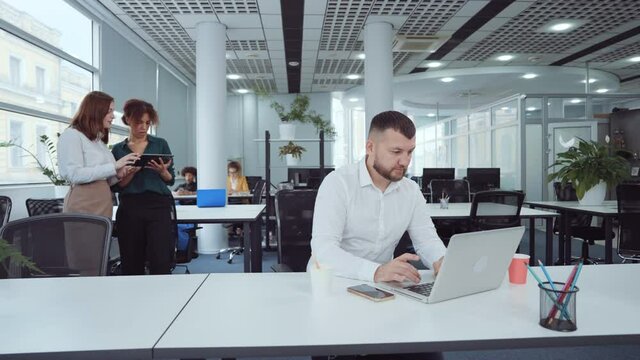 Bearded man wearing white shirt sitting in open space office and typing on laptop, shaking head in disagreement, diverse coworkers on background. Arc shot employees in shared working environment