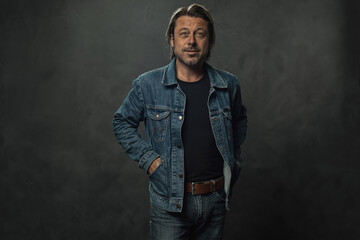 Obraz premium Blond man in jeans with a brown leather belt and a denim jacket in front of a dark grey wall.