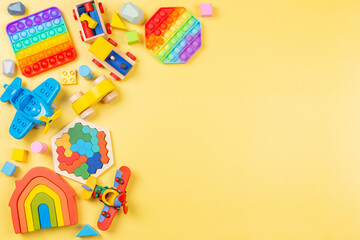 Baby kids toys background with wooden rainbow color house, train, car, plane, pop it fidget toys and colorful blocks on yellow background. Top view, flat lay