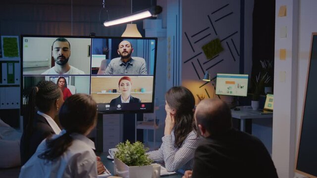 Diverse group of businesspeople discussing with remote coworkers during online videocall meeting conference. Multi ethnic teamwork working in meeting room late at night discussing company presentation