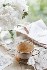 empty dirty ceramic coffee cup on creative table with scattered sheets, books, glass vase and white peonies, scattered petals. mood and atmosphere. selective focus