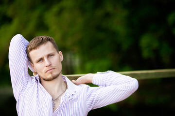 A young man with gray eyes and in a striped shirt posing on a green background from trees.