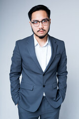 Handsome korean businessman in a suit and glasses with hands in pockets stands on a gray background