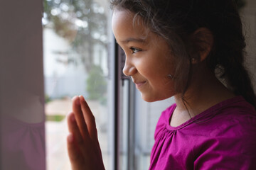 Happy hispanic girl standing at window with hand on glass, looking out and smiling