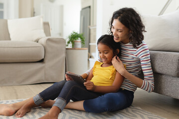 Smiling hispanic mother and daughter sitting on living room floor using tablet together