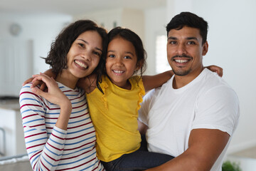 Portrait of smiling hispanic father embracing with wife and daughter standing in living room