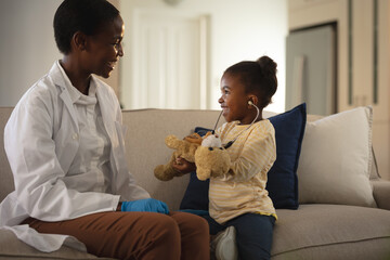 Smiling african american female doctor visiting girl patient at home, playing with stethoscope