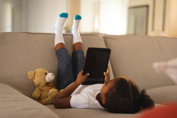 Happy african american girl upside down on couch using tablet, copy space on screen, with teddy bear