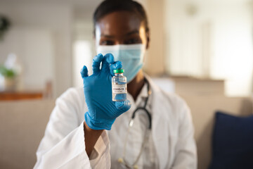 African american female doctor in face mask and gloves holding vial of covid 19 vaccine