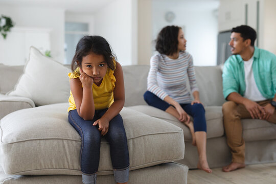 Sad hispanic daughter sitting on couch with mother and father arguing behind