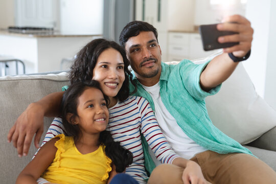 Smiling hispanic mother, father and daughter sitting on couch taking selfie together