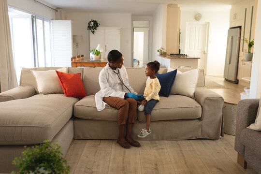 Smiling african american female doctor visiting girl patient at home, sitting on couch talking