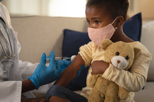 African american female doctor giving covid vaccination to girl patient in face mask at home