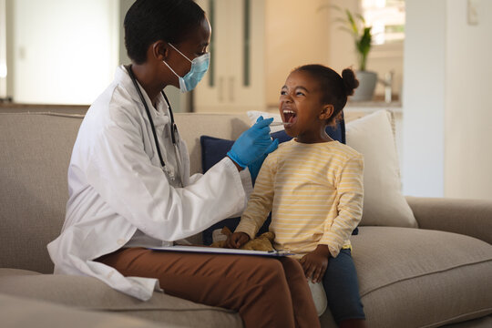African american female doctor in face mask giving girl patient covid swab test at home