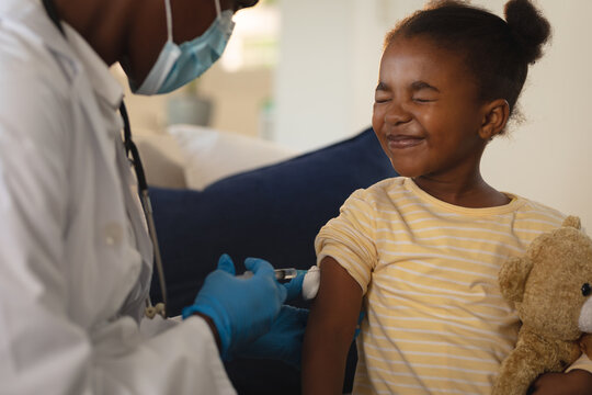 African american female doctor in face mask giving covid vaccination to scared girl patient at home