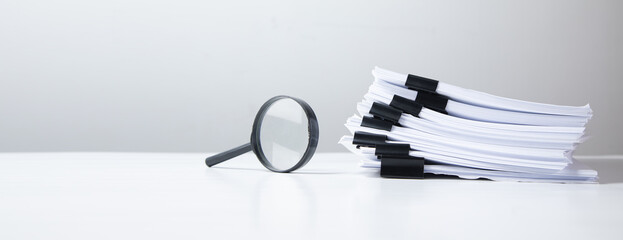 Stack of report paper documents with magnifying glass. - obrazy, fototapety, plakaty