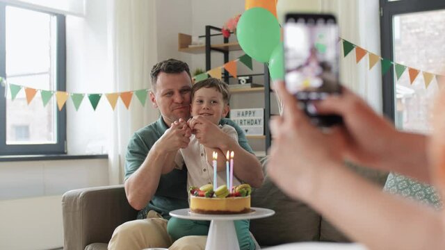 family, holidays and people concept - mother with smartphone photographing happy father and little son with birthday cake sitting on sofa at home party