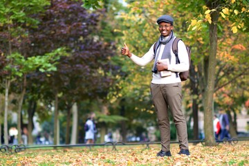 Portrait of handsome happy young black African Afro American college or university student guy with backpack and book is smiling and showing thumb up, like gesture, walking in golden autumn park. 