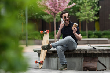 happy man in sunglasses holding paper cup and talking on smartphone near longboard and laptop.