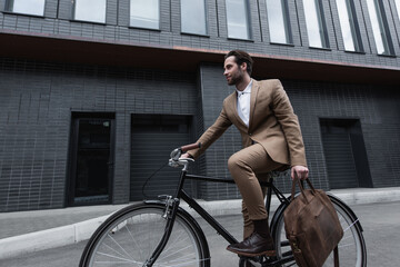 young businessman in formal wear holding leather brown bag and riding bicycle outside. - Powered by Adobe
