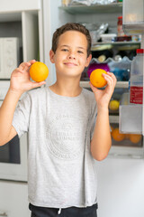 Cute healthy boy holding two orange fruits in the kitchen.