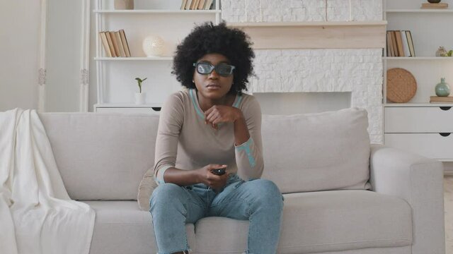 Happy young African American woman wearing 3d glasses sitting on cozy couch, switching channels using remote controller, choosing programs or shows, smiling enjoying weekend at home, leisure pastime