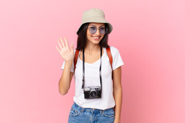 pretty hispanic tourist smiling happily, waving hand, welcoming and greeting you with a photo camera and a hat © kues1